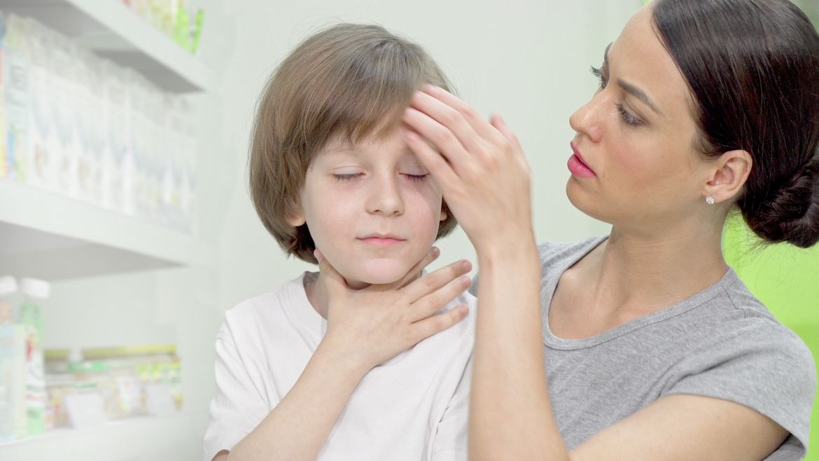 Fiebererkranktes und von Halsschmerzen geplagtes Kind fasst sich an den Hals. Mutter legt Kind Hand auf die Stirn um Fieber zu prüfen.