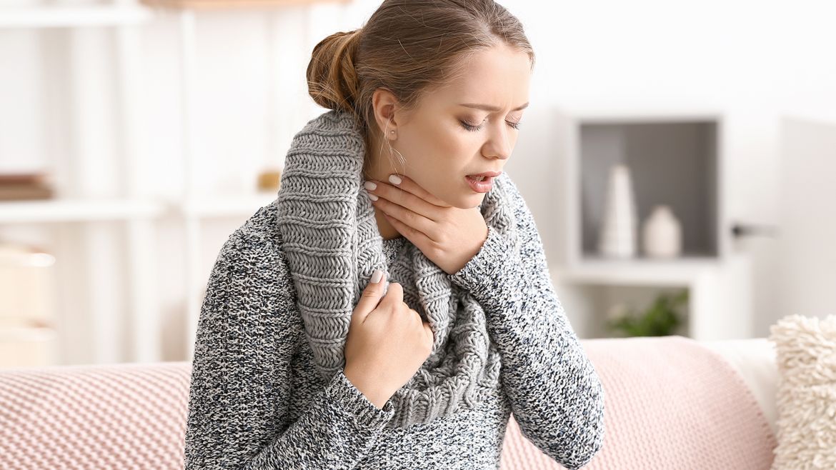 Frau mit grauem Pullover fasst sich vor Schmerzen an den Hals.