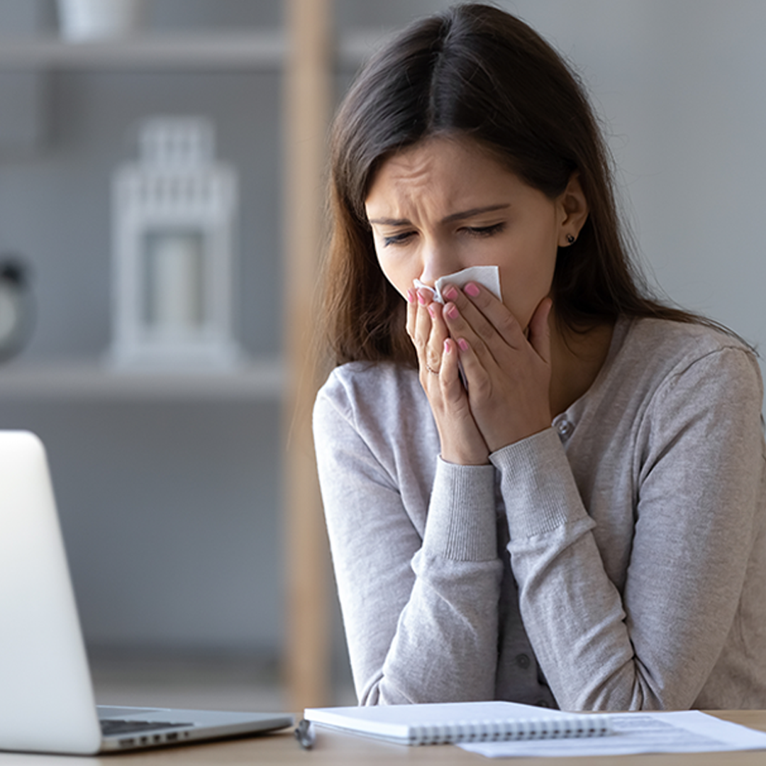 Frau sitzt vor dem Computer und ist geplagt durch Erkältungssymptome wie Schnupfen. Sie putzt sich die Nase.