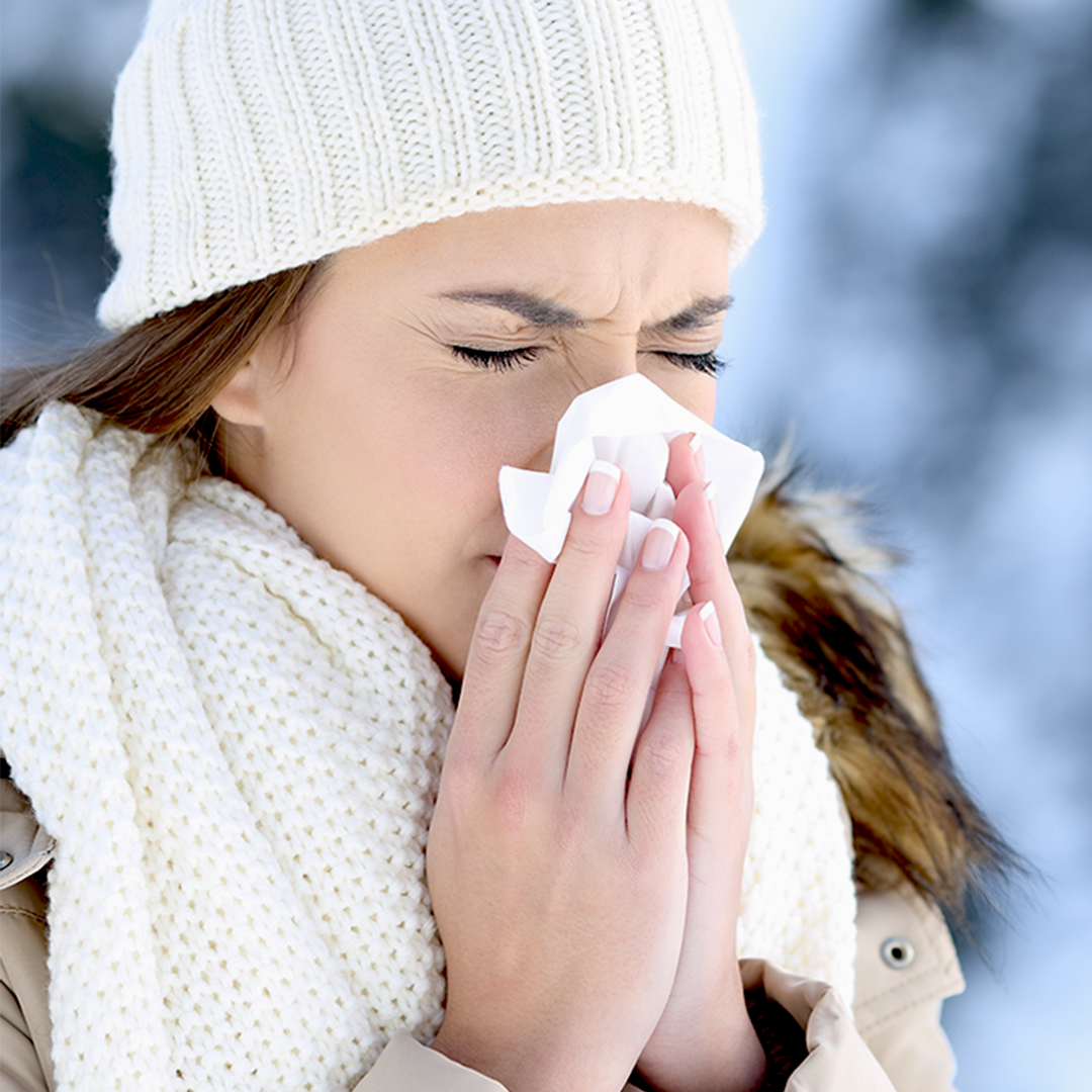 Frau mit weißer Mütze und Schal putzt sich draußen im Winter die Nase.