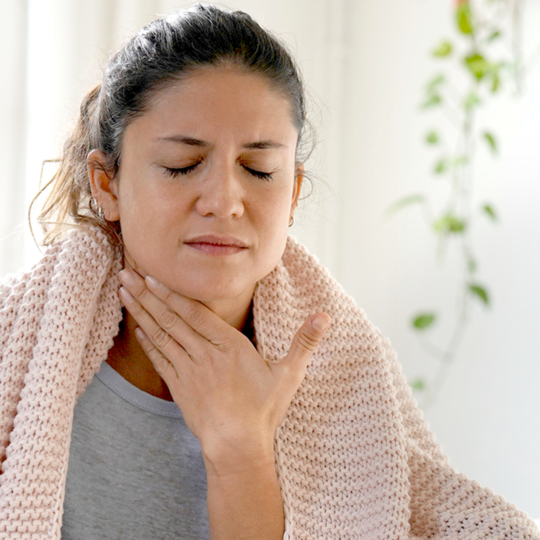 Frau ist eingewickelt in einer Decke zuhause, fasst sich durch Halsschmerzen bedingt an den Hals.